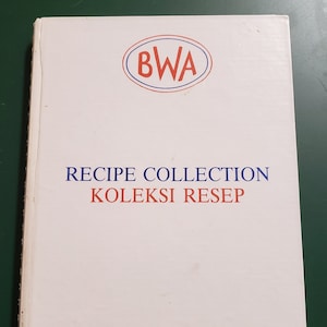 Puede incluir: Un libro blanco con un logotipo azul y rojo que dice "BWA" y el texto "RECIPE COLLECTION KOLEKSI RESEP" en rojo y azul. El libro también dice "British Women's Association" en azul en la parte inferior.