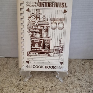 May include: A spiral-bound cookbook titled The Original Kitchener-Waterloo Oktoberfest Cook Book. The cover features a brown line-art illustration of a person cooking at a vintage stove, with pots hanging on the wall and a chair nearby.