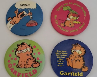 May include: Four round Garfield cartoon character pinback buttons. The buttons feature different images and text. One button has a blue background with Garfield yelling "BANZAI!" while holding a fork over a bowl of food that says "GARFIELD". Another button has a red background with Garfield holding a banana and a stalk of celery on his head. The text on this button reads "Take me to your leader... Or I'll atomize your face!" A third button has a green background with Garfield sitting with a happy expression and stars around him. The text on this button reads "GARFIELD". The final button has a blue background with Garfield sitting with a thoughtful expression. The text on this button reads "Ever have one of those days when you feel like you've eaten it all? GARFIELD".