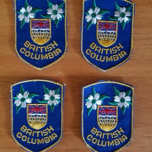 Könnte beinhalten: Vier blaue, gestickte Aufnäher in Schildform. Jeder Aufnäher zeigt weiße Blüten, ein rot-blaues Emblem und die Worte "British Columbia" in goldfarbenen Buchstaben. Die Aufnäher sind auf einer Holzoberfläche angeordnet.