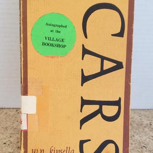 May include: A yellow book with the title "SCARS stories by w.p. kinsella" in black lettering. A green sticker with the text "Autographed at the VILLAGE BOOKSHOP" is on the front cover.
