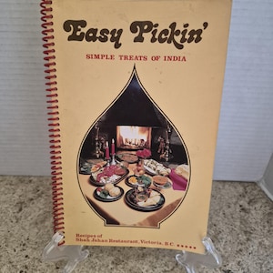 Könnte beinhalten: Ein spiralgebundenes Kochbuch mit dem Titel Easy Pickin' Simple Treats of India und einem beigefarbenen Einband. In der Mitte befindet sich ein zwiebelförmiger Ausschnitt mit einem Foto von indischen Gerichten und einem Kamin.
