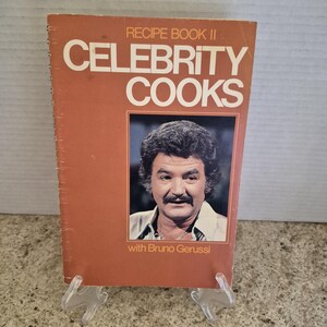 May include: A spiral-bound softcover cookbook titled Recipe Book II Celebrity Cooks with Bruno Gerussi. The cover is a solid terracotta orange colour with white text and features a rectangular photograph of a man with dark hair and a moustache.