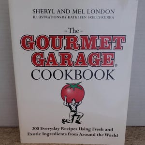 May include: The Gourmet Garage Cookbook cover features a white background with red text. The title "The Gourmet Garage Cookbook" is in large, bold red letters. A red tomato is being held up by a man in a blue shirt and khaki trousers. The text below the image reads "200 Everyday Recipes Using Fresh and Exotic Ingredients from Around the World."
