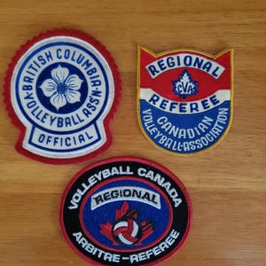 May include: Three vintage volleyball patches. The top left patch reads "British Columbia Volleyball Assn Official". The top right patch reads "Regional Referee Canadian Volleyball Association". The bottom patch reads "Volleyball Canada Regional Arbitre-Referee".