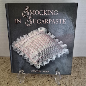 May include: A book titled "Smocking in Sugarpaste" by Cynthia Venn. The cover is dark gray with pink lettering and features an illustration of a smocked pillow with a ruffled edge. The pillow has pink and white stitching.