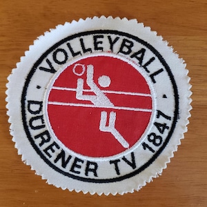 Könnte beinhalten: Eine weiße Keramik-Tasse mit schwarzem Henkel und dem Text "But First, Coffee" in fetter, schwarzer Schrift. Die Tasse steht vor einem verschwommenen Hintergrund, der ihr klares Design hervorhebt.