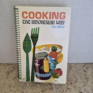 Puede incluir: Un libro de cocina con encuadernación en espiral titulado Cooking The Indonesian Way de Alec Robeau. La portada presenta un fondo blanco con texto en naranja y negro, junto a ilustraciones de un tenedor verde, una cuchara verde y varios frascos.