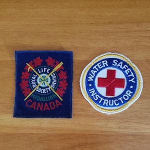 Op de afbeelding: Twee geborduurde patches op een houten oppervlak. Eén is een blauw vierkant met de tekst "ROYAL LIFE SAVING SOCIETY MEDALLION CANADA". De andere is een witte en blauwe cirkelvormige patch met de tekst "WATER SAFETY INSTRUCTOR" en een rood kruis.