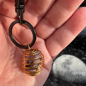 May include: A gold wire cage keychain with a pink and black stone inside. The keychain is attached to a black metal ring.