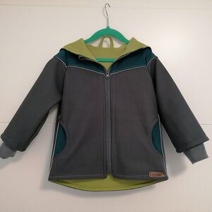 Könnte beinhalten: Eine graue Kapuzenjacke mit grünen und gelben Akzenten. Die Jacke hat einen Reißverschluss und ein kleines Etikett mit der Aufschrift "HOMEMADE".