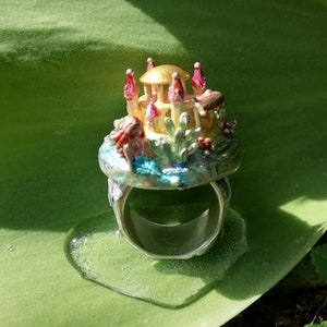 May include: A silver ring with a colorful, miniature castle-like structure on top. The castle is made of gold and pink with a green base. The ring is sitting on a green leaf.
