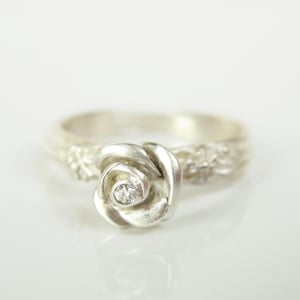 Puede incluir: Un anillo de plata con un diseño de rosa y un pequeño diamante en el centro.