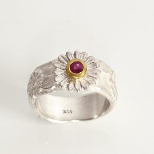 May include: A silver ring featuring a daisy design. The flower's centre is gold, set with a small, round, red gemstone. The band has a textured, organic appearance. The ring is marked with "925".
