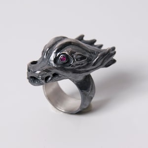 Könnte beinhalten: Ein silberner Ring mit einem detaillierten Drachenkopfdesign. Die Augen des Drachen sind mit rosafarbenen Edelsteinen besetzt.