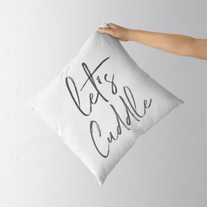 Puede incluir: Almohada blanca con texto en cursiva negra que dice "Let's Cuddle".