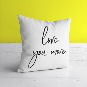 Puede incluir: Almohada blanca con texto negro que dice "love you more".