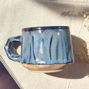 May include: A blue ceramic mug with a textured, wavy design. The mug has a wide, rounded base and a handle that curves up and out.