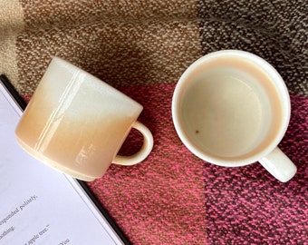 以下が含まれることがあります： 白と茶色のグラデーション釉薬をかけたセラミック製のコーヒーカップ2つ。カップは丸みを帯びた形とハンドルが付いています。