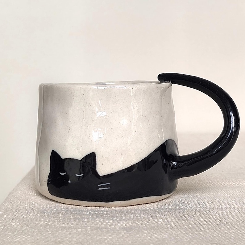 May include: A ceramic mug with a black cat design. The mug is white with a black handle and a black cat illustration wrapping around the side. The cat is lying down with its eyes closed. A unique and charming addition to any kitchen.