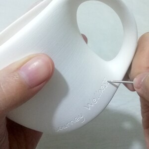 May include: A white ceramic mug with the words "Journey We're" etched into the side. A person is using a tool to etch the words into the mug.