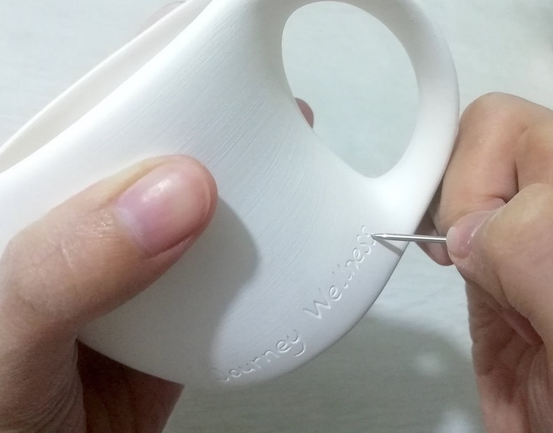 May include: A white ceramic mug with the words "Journey We're" etched into the side. A person is using a tool to etch the words into the mug.