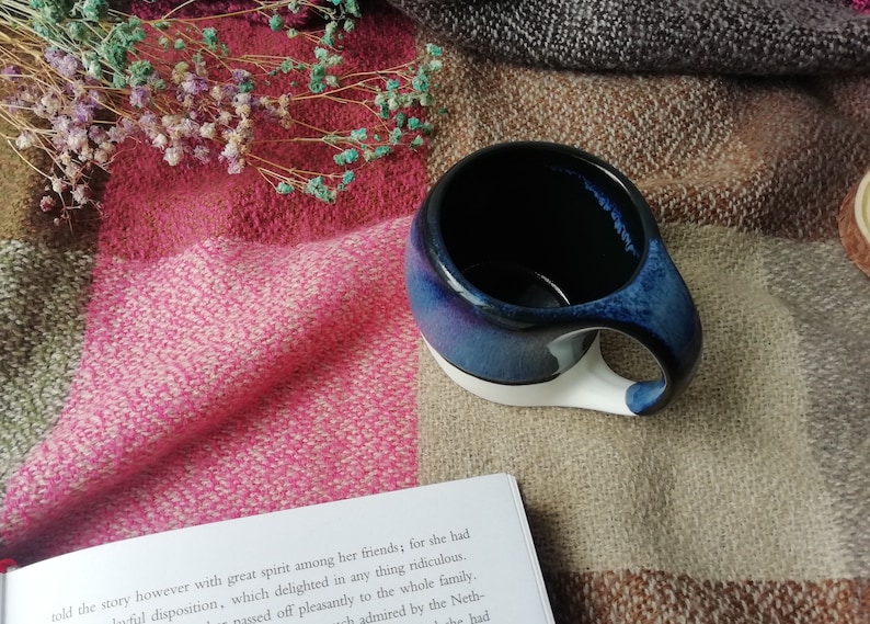 May include: A blue and white ceramic mug with a unique handle. The mug is sitting on a plaid blanket with dried flowers.