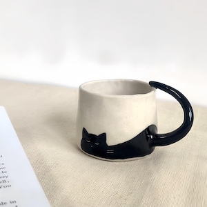 May include: A ceramic mug with a cream-colored exterior and a black cat design. The handle is a curved, dark blue. The cat is lying down, with its head and body wrapping around the mug.