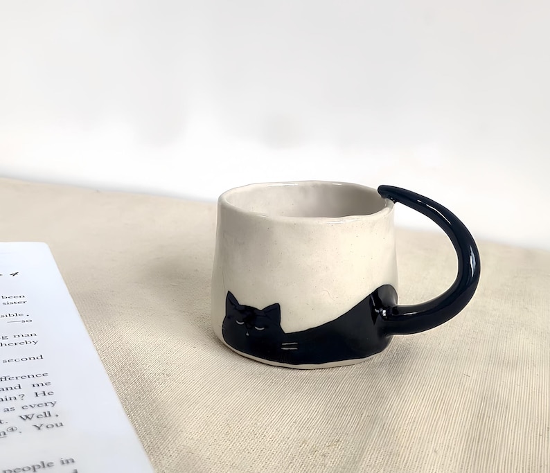 May include: A ceramic mug with a cream-colored exterior and a black cat design. The handle is a curved, dark blue. The cat is lying down, with its head and body wrapping around the mug.