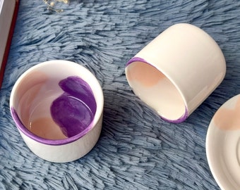 May include: Two white ceramic mugs with a purple and peach design. The mugs are on a blue fuzzy surface.