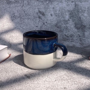 May include: A white ceramic mug with a dark blue glaze on the top half. The mug has a rounded handle.