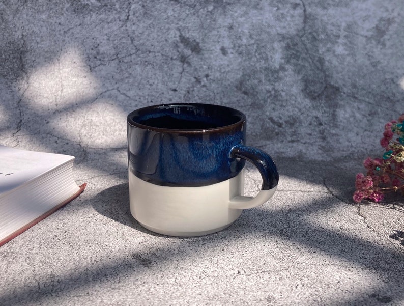 May include: A white ceramic mug with a dark blue glaze on the top half. The mug has a rounded handle.