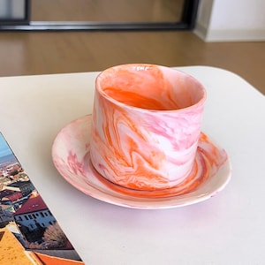Puede incluir: Un juego de taza y platillo de cerámica con un patrón arremolinado de rosa, blanco y naranja. La taza es cilíndrica y el platillo es redondo. El juego está sobre una superficie blanca.