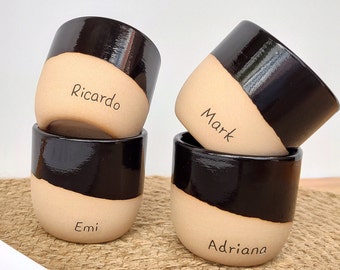 May include: Four ceramic mugs with black and beige glaze. Each mug has a different name written on the beige portion: Ricardo, Mark, Emi, and Adriana.