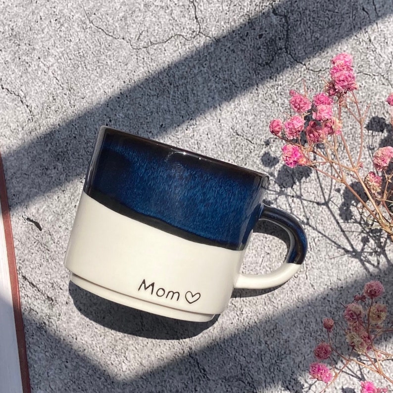 May include: A white and blue ceramic mug with a black handle. The mug has a black stripe around the top and the words "Mom" with a heart symbol written on the front.