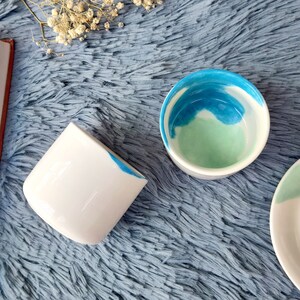 May include: Two white ceramic mugs with blue and green watercolour designs. The mugs are on a blue fluffy surface.