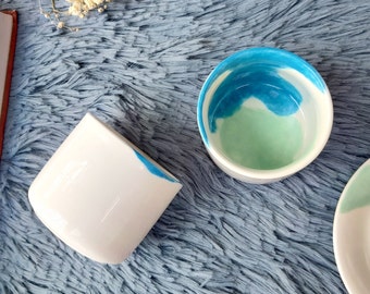 May include: Two white ceramic mugs with blue and green watercolor designs. The mugs are on a blue fuzzy surface.