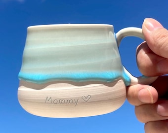 Puede incluir: Una taza de cerámica blanca con un esmalte azul claro y una línea azul ondulada en la parte inferior. La taza tiene un asa y las palabras "Mommy" y un corazón están inscritas en la parte inferior.