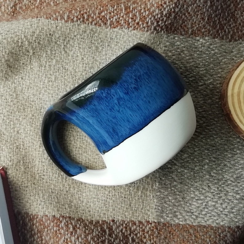 May include: A blue and white ceramic mug with a rounded shape and a handle. The mug has a dark blue glaze with a white stripe around the top.