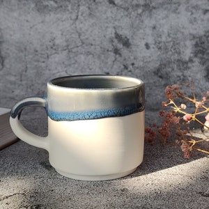 May include: A white ceramic mug with a blue and gray glaze. The mug has a rounded handle and a wide rim.