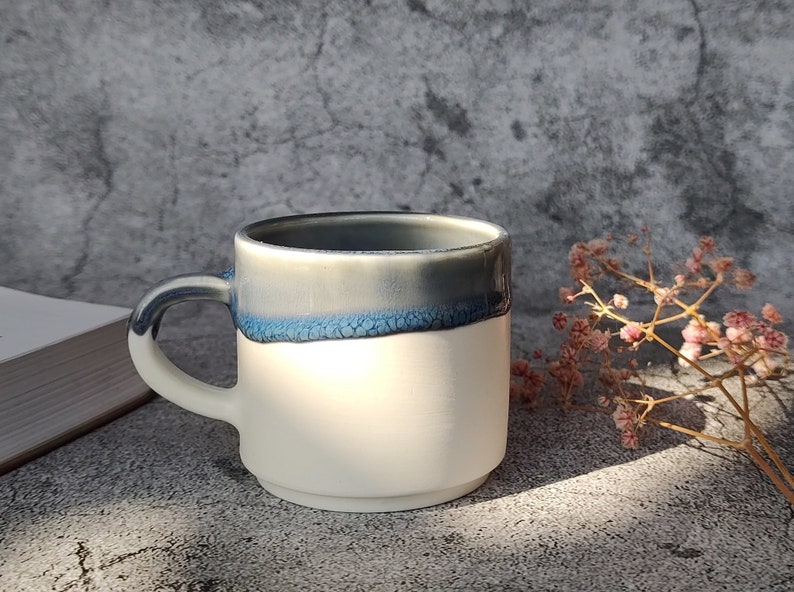 May include: A white ceramic mug with a blue and gray glaze. The mug has a rounded handle and a wide rim.