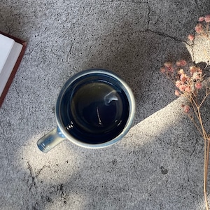 May include: A blue ceramic mug filled with dark liquid, sitting on a gray textured surface with dried flowers in the background.