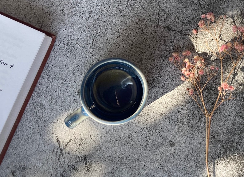 May include: A blue ceramic mug filled with dark liquid, sitting on a gray textured surface with dried flowers in the background.