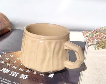 May include: A beige ceramic mug with a textured, wood-like design and a unique, sculpted handle. The mug is sitting on a magazine.