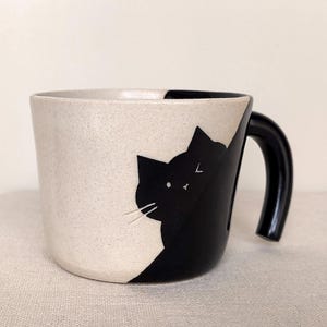 May include: A ceramic mug with a black cat design. The mug is two-toned, with a speckled beige on one side and a glossy black cat silhouette on the other. The handle is black.