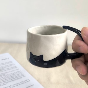 May include: A ceramic mug with a black cat design. The mug is off-white with a black handle and a black cat illustration wrapping around the side. The mug is cylindrical and appears to be handmade.