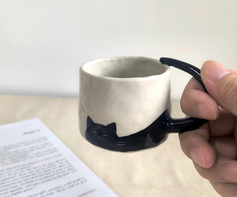 May include: A ceramic mug with a black cat design. The mug is off-white with a black handle and a black cat illustration wrapping around the side. The mug is cylindrical and appears to be handmade.