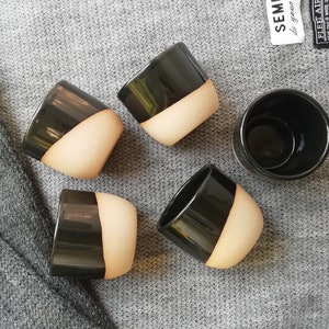 Może przedstawiać: Pięć czarnych i beżowych ceramicznych kubk&oacute;w o gładkim, zaokrąglonym kształcie. Kubki są ułożone w p&oacute;łkole na szarej dzianinowej powierzchni.