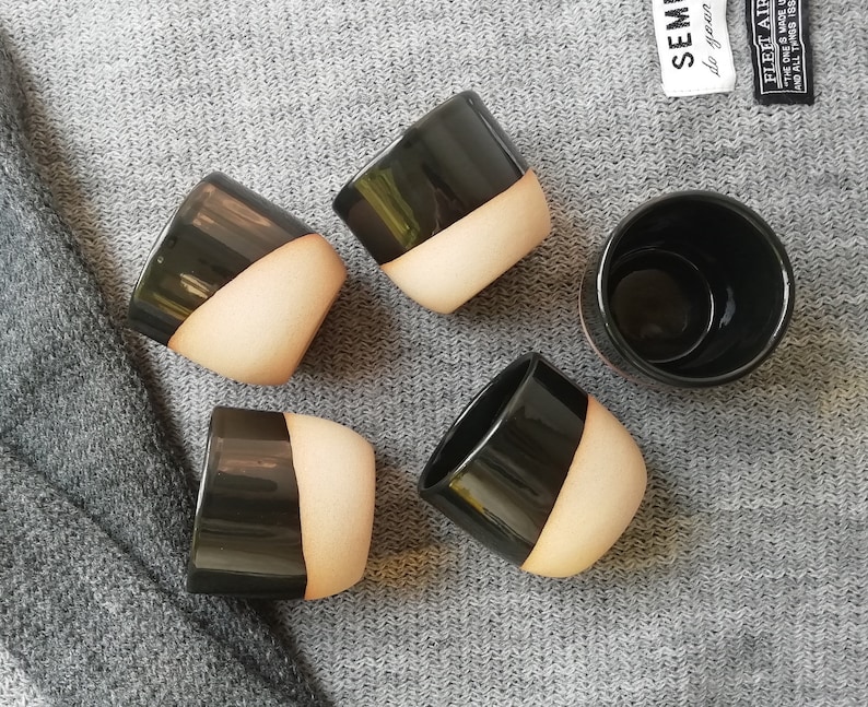 Może przedstawiać: Pięć czarnych i beżowych ceramicznych kubk&oacute;w o gładkim, zaokrąglonym kształcie. Kubki są ułożone w p&oacute;łkole na szarej dzianinowej powierzchni.
