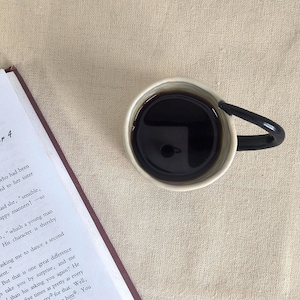 May include: A cup of dark coffee with a black handle sits next to an open book on a beige linen surface. The book is open to Chapter 4. The coffee cup is white with a black handle.